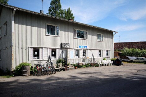 Aurinkoisena päivänä otettu valokuva vaaleasta puurakennuksesta, jonka seinässä lukee kyltissä 