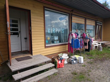 Kuvassa Kirppis Vaahteran ovensuuta ja julkisivua. Kuvasta kiitos Harrille. Valokuva pihasta, jossa vasemmalla avoinna oleva ovi ja oikealla erilaisia kirppistavaroita esillä.