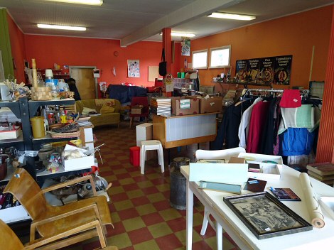 Kuvassa Kirppis Vaahteran liiketiloja. Kuvasta kiitos Harrille. Valokuva salista, jossa oranssit ja punertavat seinät. Salissa on erilaisia huonekaluja, vaatteita ja muita kirppistavaroita.
