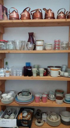 Valokuva seinää vasten seisovasta hyllyköstä täynnä erilaisia astioita ja kuparipannuja.