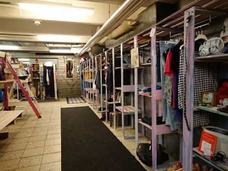 Valokuva myyntitila, jossa oikealla korkeita hyllyköitä täynnä erilaisia tavaroita. Lattialla pitkä, musta matto.