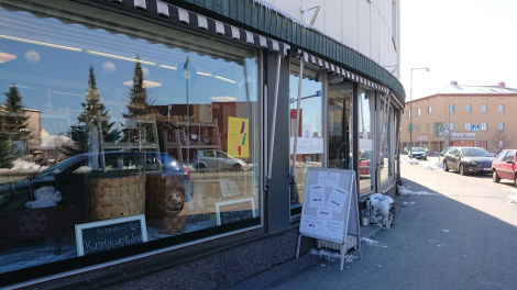 Värivalokuva Kirpputori Kuninkaankulman julkisivusta aurinkoisena päivänä. Suuret lasi-ikkunat vasemmalla puolen. Oikealla on katu, johon pysäköity henkilöautoja.
