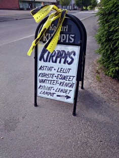 Kellarikirppiksen mainoskyltissä kerrotaan mitä sisällä odottaa. Kellarikirppiksen mainoskyltti seisoo tien vieressä.