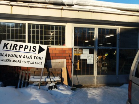 Valokuva Äijätorin sisäänkäynnistä, jossa vasemmalla suuri kyltti. Siinä lukee kirppiksen nimen alla aukioloajat.