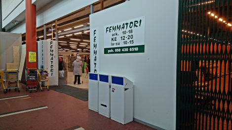 Värivalokuva Femmatorin ovensuusta. Oikealla puolen on logon alla aukioloajat ja puhelinnumero, sekä kolme roskista. Ovensuun vasemmalla puolen on kärryjä.