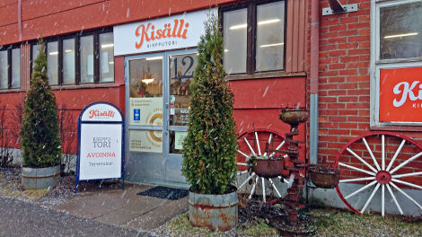 Värivalokuva punatiilisestä rakennuksen seinästä, jossa ovensuun yllä valkoista taustaa vasten Kisälli-kirppiksen logo. Ovensuun vasemmalla puolen mainosjalusta ja kummallakin puolen kaksi pientä koristepuuta.
