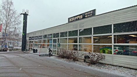 Lumiseen aikaan otettu värivalokuva kiinteistöstä, jossa seinän leveydeltä suorakulmion muotoisia ikkunoita. Katolla on suuri kyltti, jossa lukee KIRPPIS ja aukioloajat.