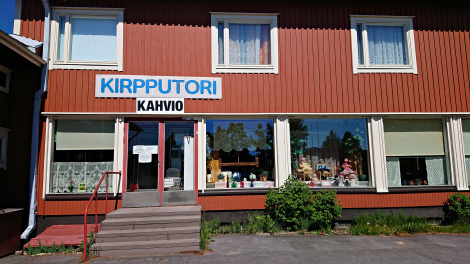 Värivalokuva punavalkoisen rakennuksen julkisivusta. Sen seinässä lukee tikkukirjaimin sana "kirpputori" ja sen alla pienemmällä "kahvio". Ovensuussa portaat, jossa neljä askelta.
