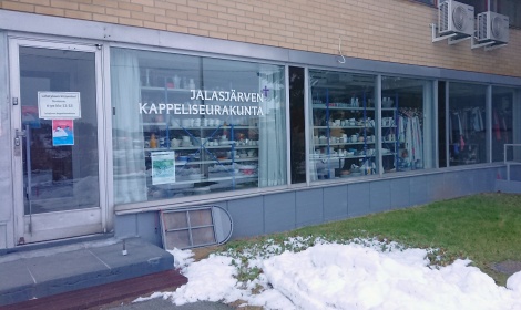 Värivalokuva Jalasjärven Kappeliseurakunnan toimipaikan julkisivusta. Vasemmalla sisäänkäynti, johon on yksi askelma, ja sen oikealla puolen suuria seinän kokoisia lasi-ikkunoita. Edessä hieman lunta vihreällä nurmikolla.