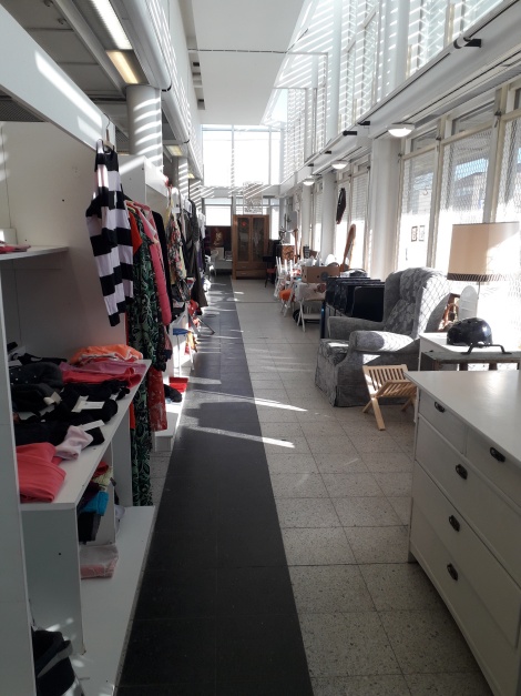 Värivalokuva pitkästä käytävästä, jossa oikealla puolen seinän pituudelta suuria ikkuinoita ja vasemmalla valkoisia kirppishyllykköjä täynnä värikkäitä tavaroita.