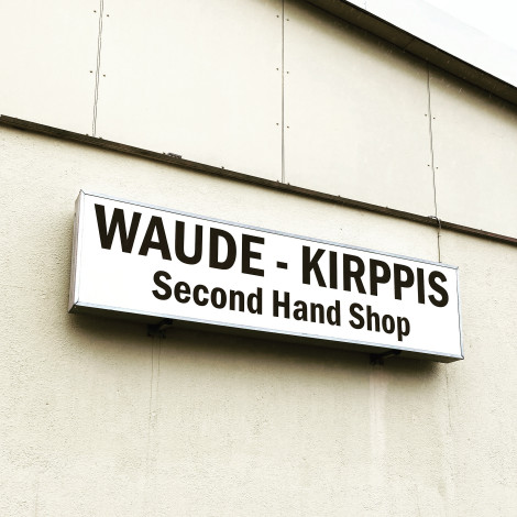 Värivalokuva, jossa valkoiselle seinälle ripustettu mainoskyltti. Siinä lukee mustalla värillä "Waude-kirppis", ja sen alla "Second Hand Shop".