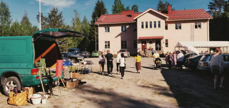 Värivalokuva kaksikerroksisesta talosta, jonka edessä hiekkakenttä täynnä ihmisiä ja peräkonttikirppiksen pitäjiä.