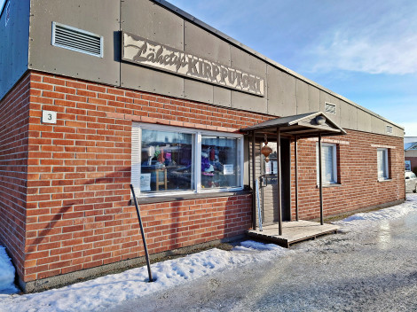 Talvipäivänä otettu värivalokuva yksikerroksisesta tiilitalosta, jossa tiilet ovat punertavia. Katon yläraja on tummanharmaa ja siinä on koristeellinen Lähetyskirpputorin liikekyltti.