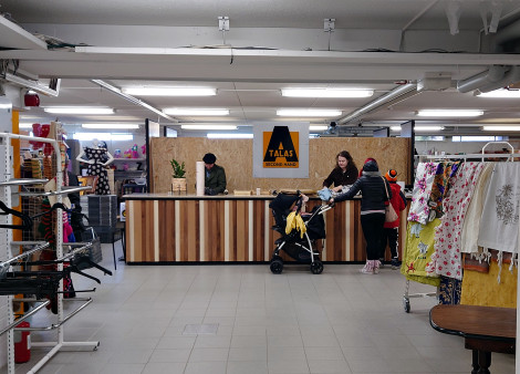 Kuvassa värivalokuva kassa-alueesta, jonka ympärillä hyllyjä ja rekkejä täynnä erilaisia esineitä, kuten vaatteita ja kankaita.