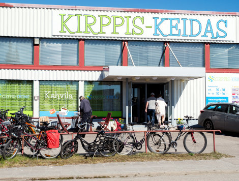 Värivalokuva liikekiinteistöstä, jonka seinällä lukee suurin kirjaimin "Kirppis-Keidas". Sen alla on ovensuu. Etualalla on polkupyöriä ja taustalla viipale sinistä taivasta.