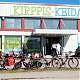 Värivalokuva liikekiinteistöstä, jonka seinällä lukee suurin kirjaimin "Kirppis-Keidas". Sen alla on ovensuu. Etualalla on polkupyöriä ja taustalla viipale sinistä taivasta.