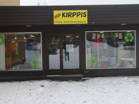 Talvisena päivänä otettu värivalokuva kiinteistön julkisivusta. Edessä suuret lasi-ikkunat ja niiden välissä sisäänkäynti, joka sekin on lasiovi. Sisäänkäynnin yläpuolella mainoskyltti, jossa lukee HYMIS-Kirppis.