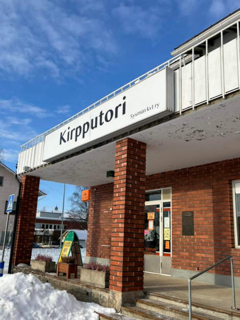 Talvisena päivänä otettu värivalokuva tiiliskivistä rakennetusta kiinteistöstä, jonka juurella on portaat, vasemmalla lumikasa, ja seinällä suuri kyltti, jossa lukee: Kirpputori Sysmän kvt ry.