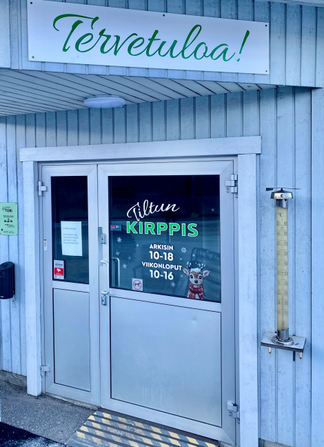 Värivalokuva, jossa näkyy sininen puuseinä ja ovi, jossa on Tiltun Kirppiksen logo. Oven yläpuolella on kyltti. Siinä lukee: Tervetuloa!
