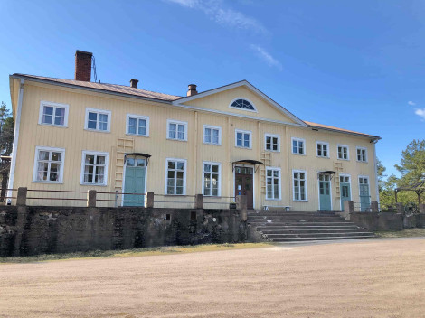 Värivalokuva suuresta puurakennuksesta, jossa keltavalkoiset seinät. Etualalla hiekkapiha ja talolle johtavat valtavat kiviportaat.