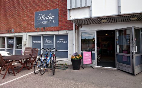 Kuvassa Hiiden Kirppiksen ovensuu. Hiiden Kirppiksen julkisivu: ovet auki ja vasemmalla puolen pyöräteline sekä pöytä tuolien kanssa.