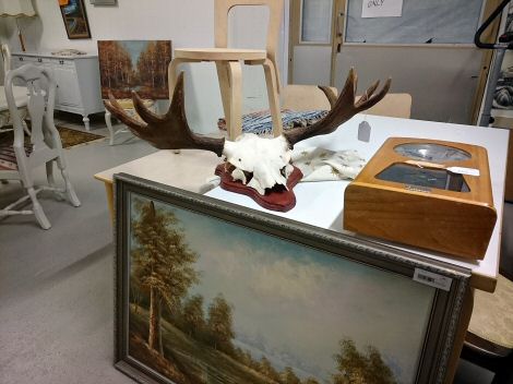 Yksityiskohtia Vintikki-kirppiksen tavaravalikoimasta. Suuri maisemataulu pöytää vasten ja pöydän päällä metsästystrofee.